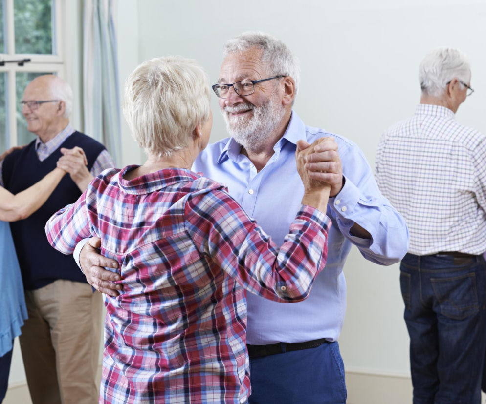 Szczęśliwi seniorzy tańczący w parach w jasnym pomieszczeniu, z oknem w tle. Uśmiechnięte twarze i wspólna aktywność fizyczna sugerują radość z życia i dbałość o sprawność umysłu, o czym więcej przeczytasz na Poradniku Zdrowie.