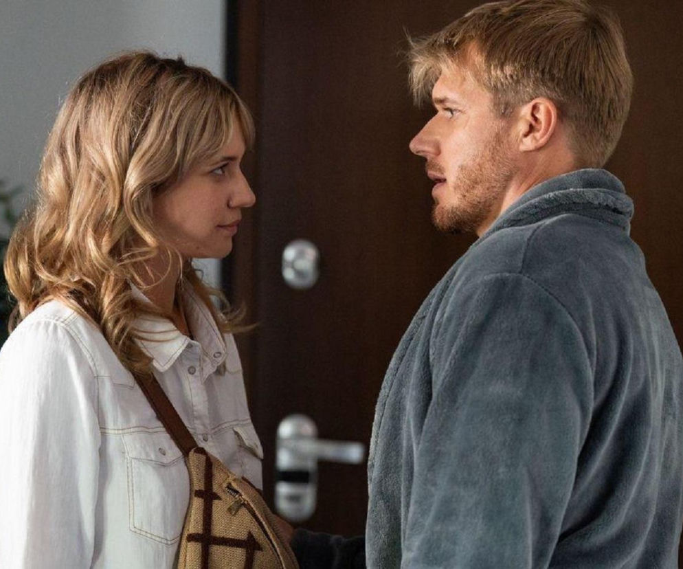 Gloria (Aleksandra Grabowska) i Maks (Tomasz Ciachorowski) stoją naprzeciwko siebie w pokoju. Gloria, w białej koszuli, patrzy na Maksa, który w szarym szlafroku ma otwarte usta, jakby mówił. Dramatyczna scena z serialu Na dobre i na złe dostępna na Super Seriale.