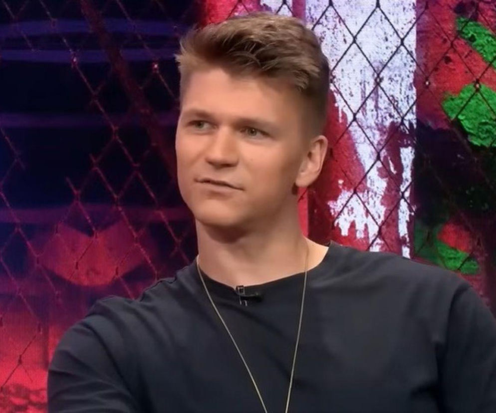 Komik Roman Kostelecki w ciemnym t-shircie na tle siatki ogrodzeniowej. O jego pracy w Polsce przeczytasz na Mediateka.