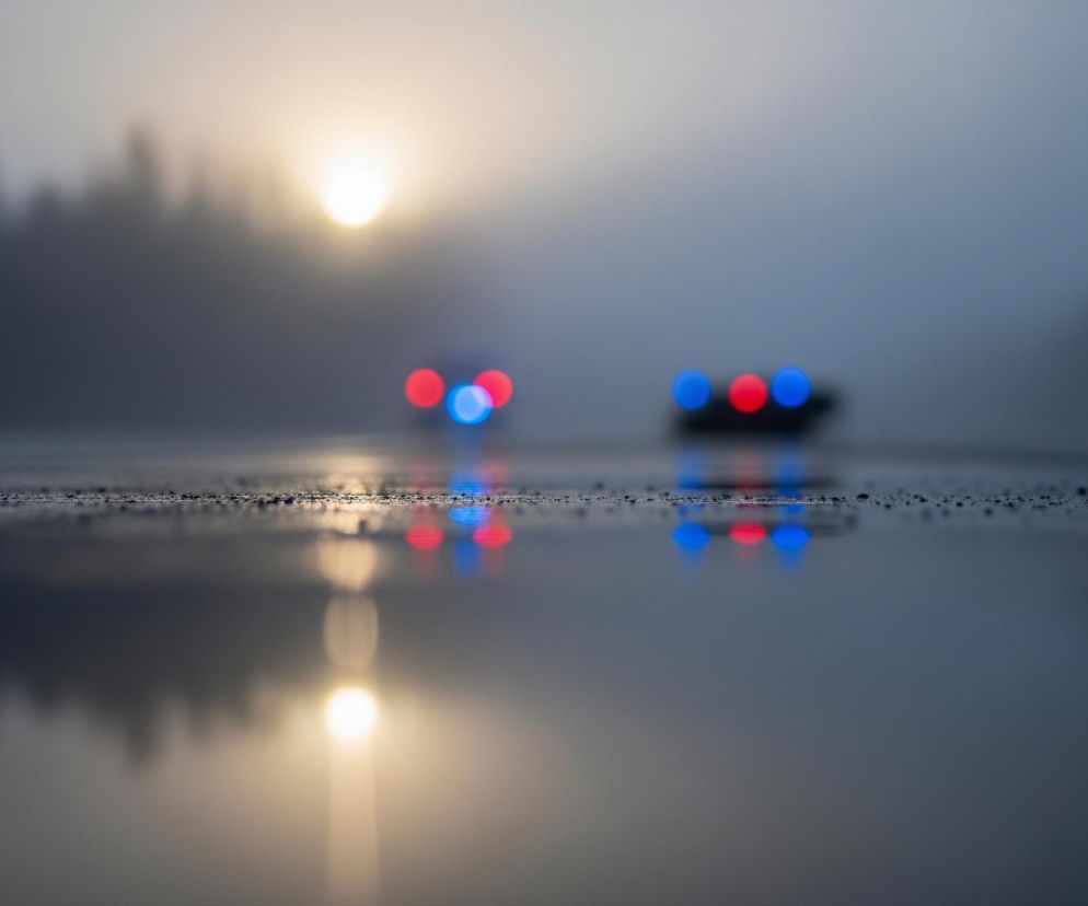 Na pierwszym planie widoczna jest mokra nawierzchnia pokryta drobnymi kroplami wody, odbijająca otoczenie jak lustro. W oddali, w tle, wyróżniają się dwa zamglone kształty samochodów z wyraźnie widocznymi, rozmazanymi światłami w kolorach czerwonym i niebieskim. Nad horyzontem, po lewej stronie obrazu, widać jasne, rozmyte słońce, które przebija się przez mgłę, tworząc poświatę. Cała scena jest zdominowana przez odcienie szarości, błękitu i ciemnych tonów, z wyjątkiem intensywnych barw świateł i słońca.