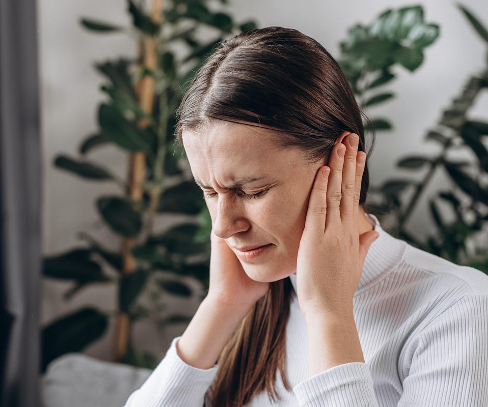 Kobieta w białej bluzce z długimi, ciemnymi włosami, trzymająca dłonie przy uszach i mająca zmarszczone brwi, co symbolizuje odczuwany ból lub dyskomfort związany z szumami usznymi. Tło jest rozmyte, widoczne są liście rośliny doniczkowej. Poradnik Zdrowie oferuje informacje, jak radzić sobie z tym problemem.