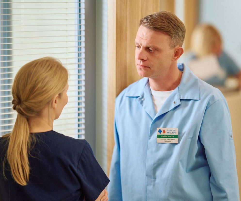 Doktor Kamil Hoffer w błękitnym kitlu szpitalnym z plakietką Szpital Miejski, Kardiologia, dr n. med. Kamil Hoffer, Ordynator rozmawia z koleżanką po fachu w ciemnoniebieskim uniformie, stojąc w korytarzu szpitalnym obok okna z żaluzjami. Więcej o losach bohaterów serialu Na Wspólnej na Super Seriale.