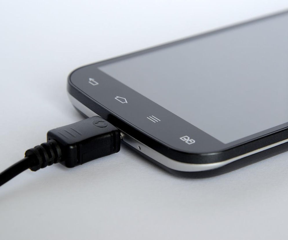 Czarny smartfon podłączony do kabla ładowarki. O kosztach ładowania telefonu w skali roku przeczytasz na SE Superbiz.