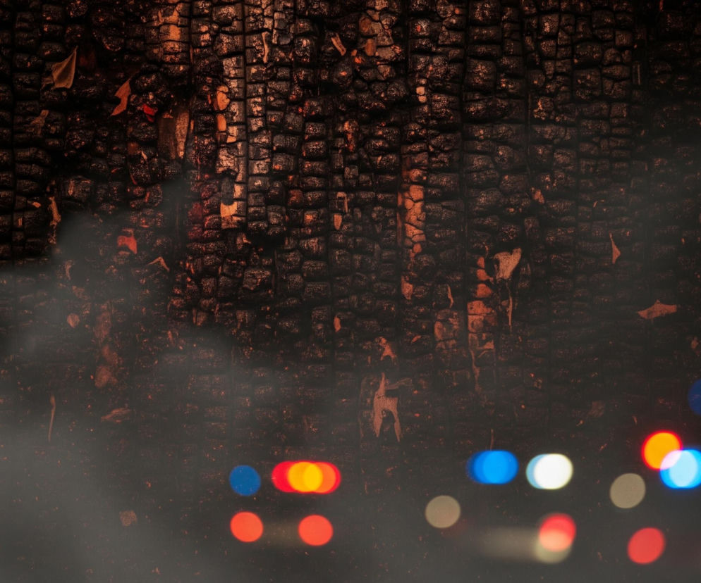 Górna część obrazu przedstawia teksturę spalonego drewna, z nieregularnymi, ciemnymi fragmentami i jaśniejszymi, pomarańczowymi akcentami widocznymi w szczelinach. Dolna połowa sceny jest pokryta szarym dymem lub mgłą, przez którą przebijają się rozmazane światła w kolorach czerwonym, pomarańczowym, niebieskim i białym, ułożone w grupy, przypominające lampy pojazdów. Całość utrzymana jest w ciemnej tonacji z dominacją czerni i brązu, przełamaną jaskrawymi plamami świateł.