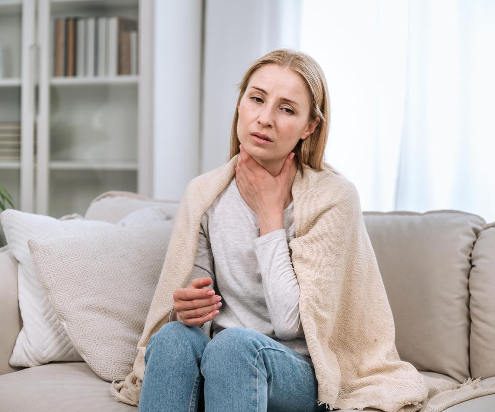 Kobieta z objawami bólu gardła, siedząca na kanapie z kocem, trzymająca dłoń na szyi. Obraz symbolizuje dolegliwości tarczycy i potrzebę diagnostyki, o której można przeczytać na portalu Poradnik Zdrowie.