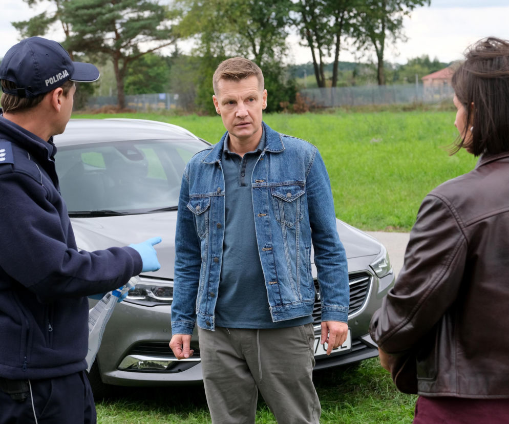 Paweł Zduński (Rafał Mroczek) w dżinsowej kurtce rozmawia z policjantem i Agnes (Amanda Mincewicz) na tle szarego samochodu, po incydencie w Grabinie. Zdjęcie ilustruje scenę z serialu M jak miłość, o której można przeczytać na portalu Super Seriale.