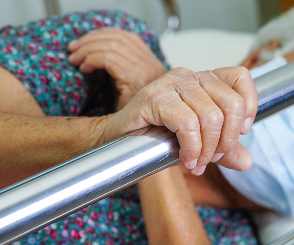 Dłoń seniorki trzymająca metalową barierkę łóżka. O kryzysie w opiece nad starszymi osobami przeczytasz na SE Superbiz.