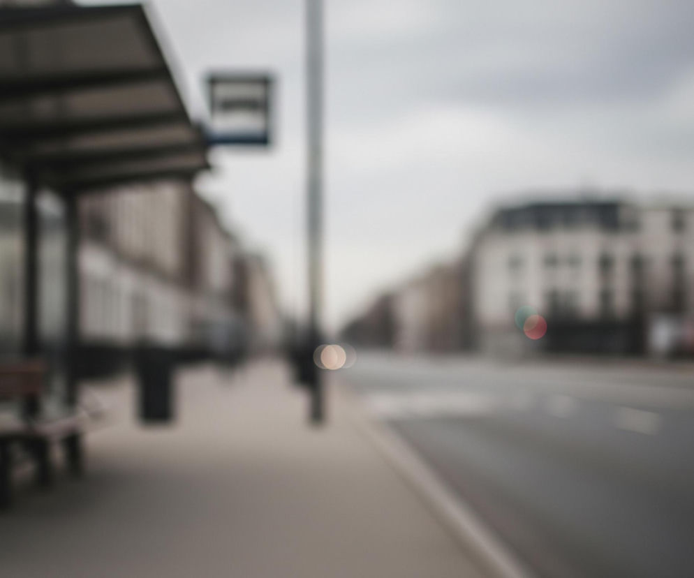 Rozmyty obraz przedstawia ulicę miejską z przystankiem autobusowym po lewej stronie. W pierwszym planie znajduje się metalowa konstrukcja przystanku w ciemnych kolorach, z częściowo widocznym ogłoszeniem i drewnianą ławką. Po prawej stronie biegnie szara jezdnia z białymi, rozmytymi liniami poziomymi, a w oddali widać niewyraźne budynki i drzewa pod jasnym, zachmurzonym niebem.