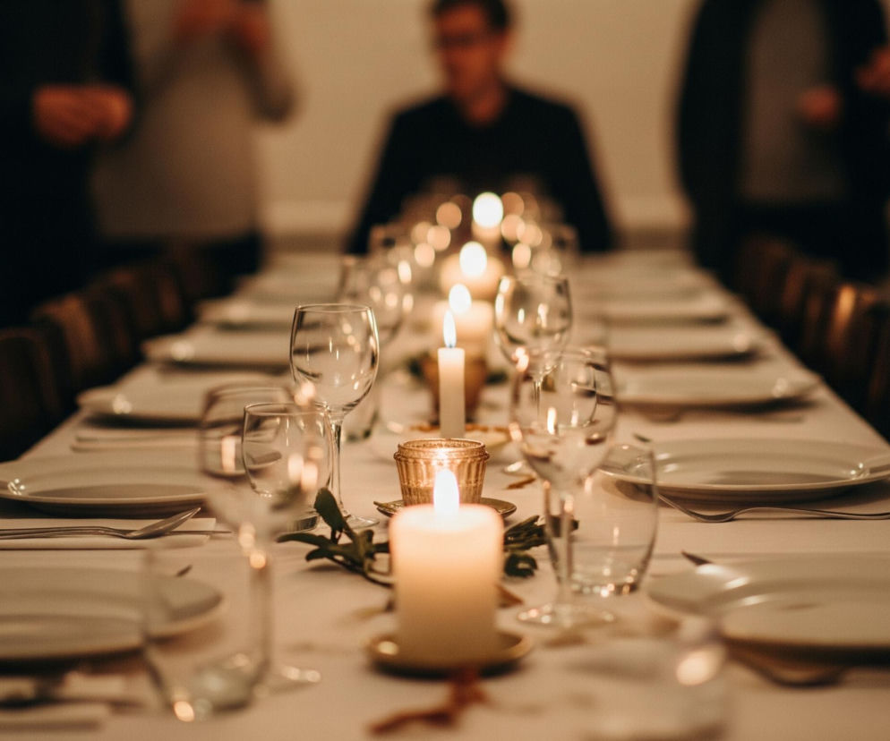 Długi stół pokryty białym obrusem jest centralnym punktem obrazu, zastawiony rzędami białych talerzy, srebrnych sztućców i przezroczystych kieliszków, rozmieszczonych po obu stronach. Wzdłuż stołu stoją zapalone świece o ciepłym, żółtym płomieniu, niektóre w szklanych świecznikach, inne bezpośrednio na złotych podstawkach, z zielonymi gałązkami ułożonymi pomiędzy nimi. W tle, niewyraźne sylwetki ludzi w ciemnych ubraniach stoją lub siedzą, oświetlone jedynie delikatnym blaskiem świec, tworząc ciepłą i intymną atmosferę.