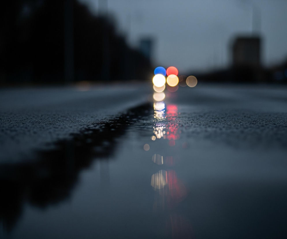 Mokry asfalt uliczny, pokryty drobnymi kroplami wody, zajmuje pierwszy plan i rozciąga się w głąb obrazu. Na horyzoncie widoczne są rozmyte światła samochodów: cztery wyraźne, okrągłe punkty, w tym jeden niebieski, jeden czerwony oraz dwa żółte, z towarzyszącymi im mniejszymi, jaśniejszymi światłami. Ich odbicia, mniej ostre, lecz z wyraźnie zaznaczonymi kolorami, padają na mokrą nawierzchnię, tworząc pionowe pasy świetlne. Tło stanowi ciemny, rozmyty pejzaż miejski z niewyraźnymi sylwetkami budynków i drzew.