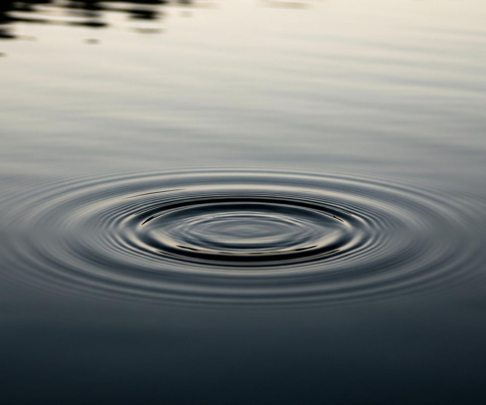 Na pierwszym planie, centralnie, widoczne są koncentryczne kręgi na spokojnej tafli wody. Najjaśniejszy punkt znajduje się w samym środku okręgów, od którego rozchodzą się fale o zmiennej intensywności barwy, od ciemnego granatu w zagłębieniach po jaśniejsze, prawie kremowe odcienie na szczytach fal. Powierzchnia wody w dolnej części obrazu jest ciemnogranatowa, niemal czarna, przechodząc stopniowo w jaśniejszy, szarobeżowy odcień ku górze. W lewym górnym rogu widać rozmyte, ciemne plamy, które mogą być odbiciem obiektów znajdujących się poza kadrem.