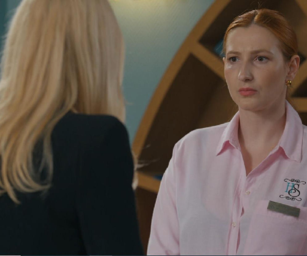 Aktorka Anna Maria Kosik jako Sylwia, ubrana w różową koszulę z logo i identyfikatorem, patrzy na rozmówczynię w czarnym żakiecie. Scena z serialu „Barwy szczęścia”, gdzie Sylwia ma zastąpić Kajtka w hotelu. Więcej o losach bohaterów przeczytasz na Super Seriale.