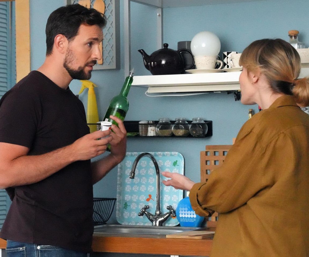 Marcin Kodur (Oskar Stoczyński) trzyma butelkę i opakowanie leków, rozmawiając z Renatą (Anna Mrozowska), która ma na sobie musztardową koszulę, w kuchni serialu Barwy szczęścia. Więcej o tym na portalu Super Seriale.