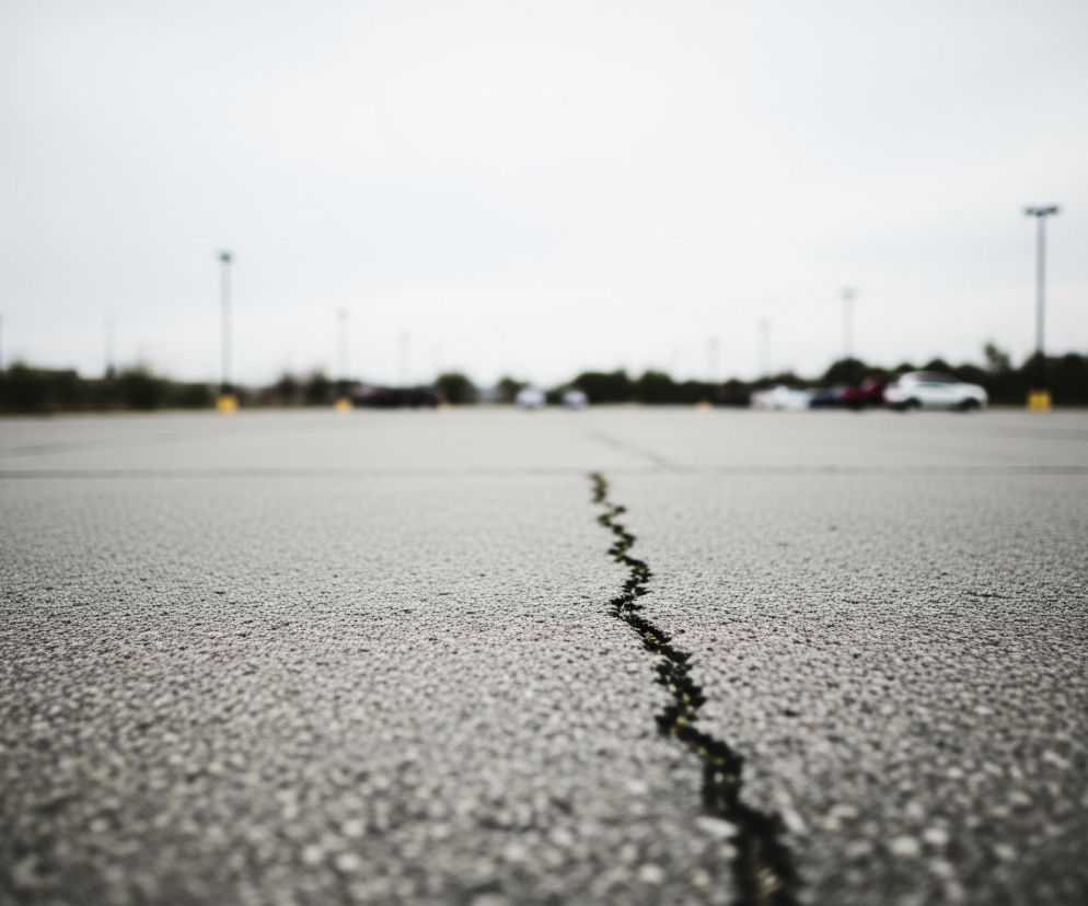 Na pierwszym planie widoczna jest szara, asfaltowa nawierzchnia z wyraźnym, zygzakowatym pęknięciem biegnącym od dołu kadru w kierunku środka, wypełnionym zielonkawymi elementami. Tło, znacznie rozmyte, przedstawia szeroki parking lub plac, na którym w oddali stoją niewyraźne sylwetki samochodów i wysokie, ciemne słupy oświetleniowe, niektóre z żółtymi podstawami. Horyzont wyznacza linia ciemnych krzewów i drzew, a nad nimi rozciąga się jasne, zachmurzone niebo.