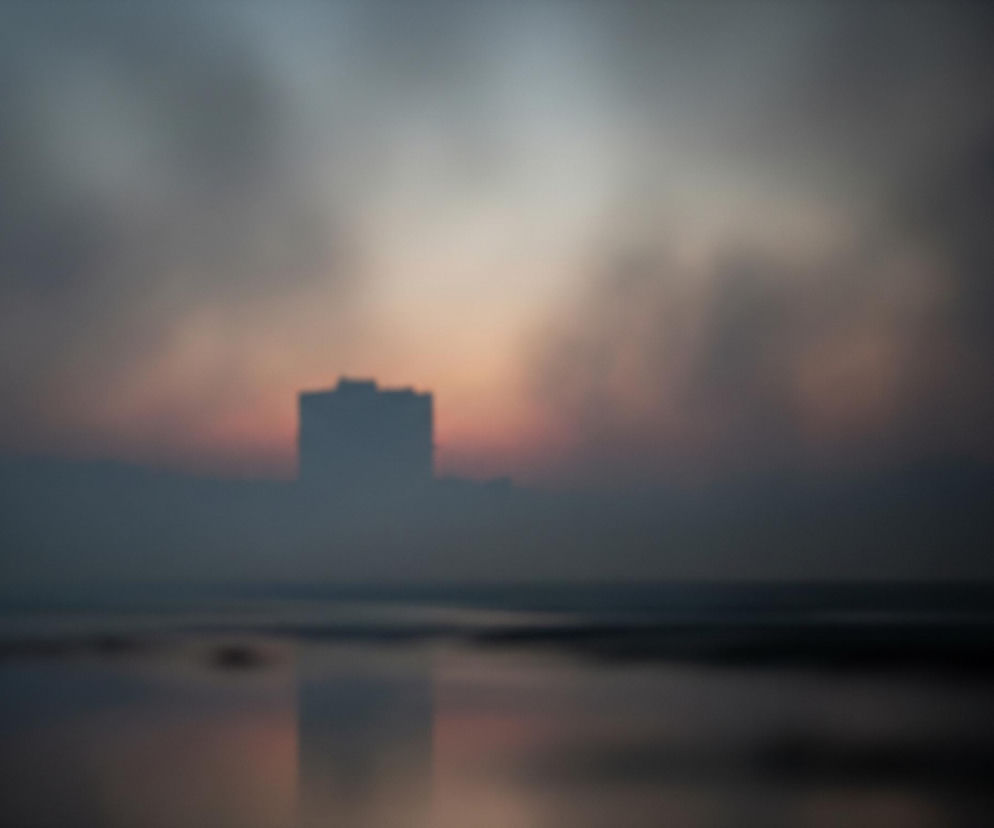 Rozmazana, ciemna sylwetka budynku, przypominająca wieżowiec, dominuje na horyzoncie, odbijając się w rozmytej, odblaskowej powierzchni na pierwszym planie. Niebo za budynkiem przybiera odcienie szarości i pomarańczowo-różowego rozbłysku, sugerując zachód słońca lub świt, wszystko otulone gęstą mgłą lub dymem. Cały obraz jest nieostry, nadając mu zamglony, eteryczny charakter, gdzie granice między niebem, budynkiem i ziemią są zatarte. Dominują chłodne barwy szarości i błękitu, przełamane ciepłymi akcentami pomarańczu i różu w centrum.