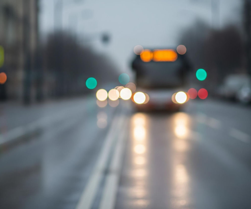 Ulica w mieście o zmierzchu lub w nocy, z widocznym w oddali rozmytym kształtem pojazdu, prawdopodobnie autobusu, z wyraźnymi pomarańczowymi światłami na górze i jaśniejszymi, okrągłymi reflektorami z przodu. Nawierzchnia drogi jest mokra, odbijając rozproszone światła latarni i innych pojazdów jako podłużne smugi i owalne plamy. Po obu stronach jezdni, wyznaczonej podwójną ciągłą linią, znajdują się rozmazane sylwetki budynków i drzew, a także szereg świateł w kształcie zielonych i czerwonych kręgów.