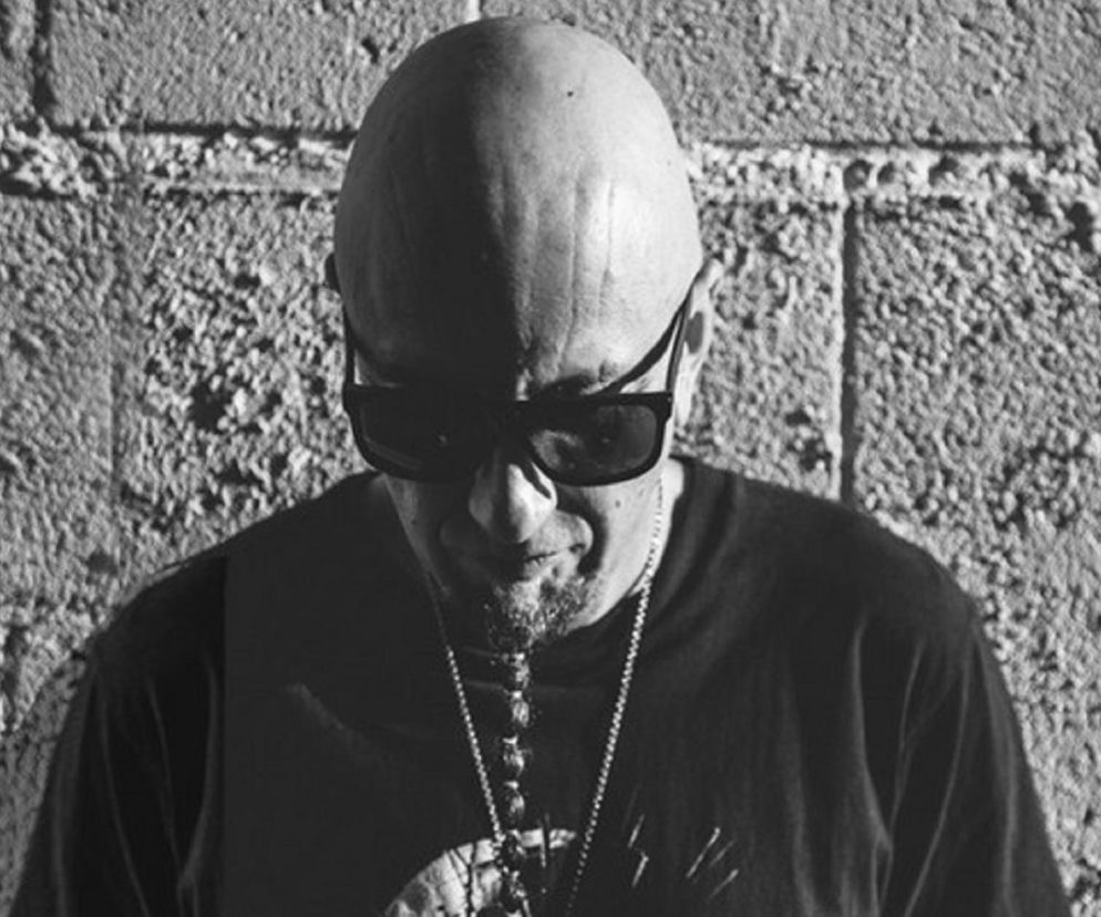 Muzyk Shavo Odadjian w okularach przeciwsłonecznych na tle muru. O jego nowej roli przeczytasz na EskaRock.