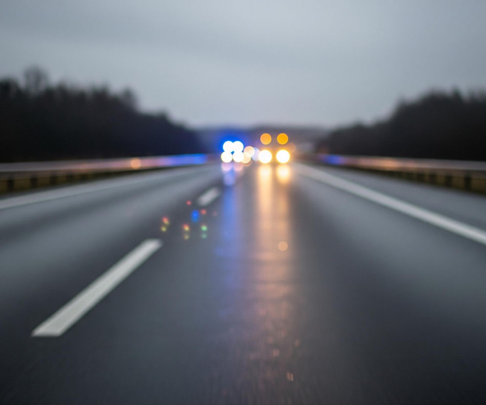 Asfaltowa droga, prawdopodobnie autostrada, rozciąga się w kierunku centrum kadru, z widocznymi dwoma pasami ruchu rozdzielonymi przerywaną białą linią po lewej stronie i ciągłą białą linią po prawej. Po obu stronach drogi biegną barierki ochronne w kolorze stalowym z żółtymi elementami u podstawy. W oddali, na horyzoncie, widać rozmyte światła samochodów: wyraźne niebieskie światła pojazdu uprzywilejowanego oraz liczne, rozmazane pomarańczowo-żółte światła innych pojazdów, tworzące odblaski na mokrej nawierzchni. Tło stanowią rozmazane, ciemne sylwetki drzew pod pochmurnym, szarym niebem, dodając zdjęciu mrocznego i wilgotnego charakteru.
