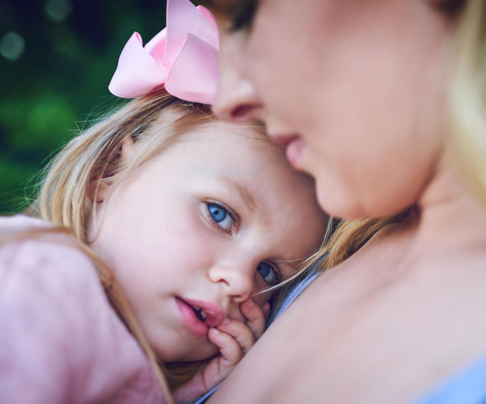 Dziewczynka z blond włosami i niebieskimi oczami, z różową kokardą, wtulona w ramię dorosłej osoby. Zdjęcie symbolizuje potrzebę bezpieczeństwa i lęku przed obcymi, o czym więcej przeczytasz na naszym portalu.