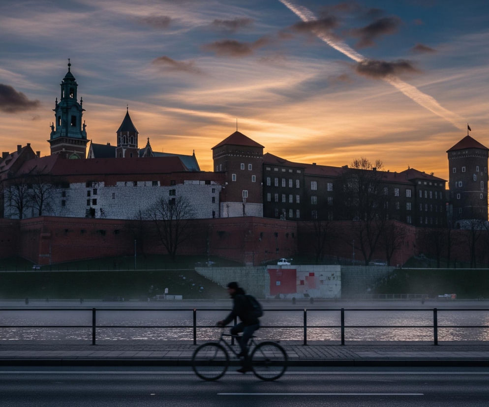 Mężczyzna na rowerze, rozmyty ruchem, jedzie w lewą stronę po ciemnym, asfaltowym chodniku biegnącym wzdłuż rzeki, oddzielonej czarną, metalową barierką od betonowej ścieżki. Za rzeką unosi się lekka mgła, a na jej drugim brzegu widoczny jest masywny kompleks budynków z ceglanymi murami i dachami, w tym wysoka wieża z iglicą. Niebo nad budowlami jest gradientem od ciemnoniebieskiego na górze do jasnopomarańczowego przy horyzoncie, z widocznymi smugami chmur i dwiema smugami kondensacyjnymi samolotów przecinającymi się.