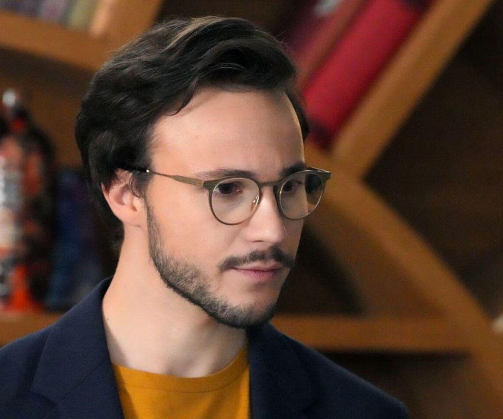 Młody mężczyzna, Jakub Dmochowski jako Kajtek w serialu „Barwy szczęścia”, z krótką brodą i okularami, ubrany w żółtą koszulkę i granatową marynarkę, patrzący w bok z zamyślonym wyrazem twarzy na tle drewnianego regału. Więcej o wątku Karoliny i Kajtka znajdziesz na Super Seriale.