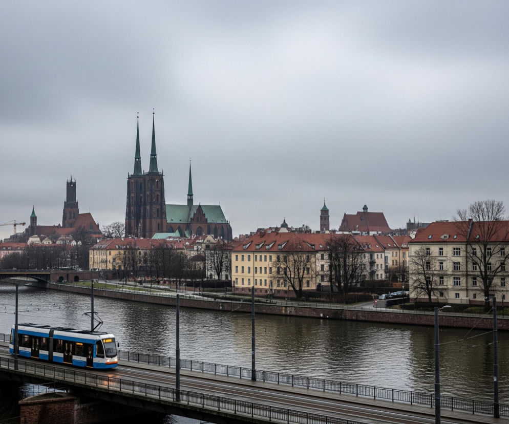 Błękitno-biały tramwaj przejeżdża po moście nad rzeką, z kilkoma osobami spacerującymi po chodniku obok torów. Na brzegu rzeki widać rząd budynków o żółtych i kremowych fasadach oraz czerwonych dachach, za którymi wznoszą się strzeliste wieże gotyckiego kościoła. Niebo jest zachmurzone, a szare, spokojne wody rzeki odzwierciedlają otoczenie.