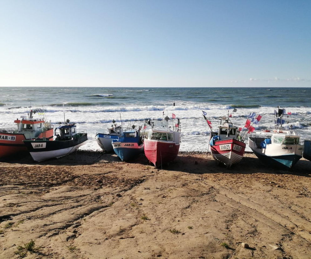 Rząd kolorowych kutrów rybackich z numerami JAR na piaszczystej plaży Jarosławca, z Bałtykiem i falami w tle. To typowy widok w miejscowości, która zyska nowoczesny punkt informacji turystycznej, o czym przeczytasz na naszym portalu.