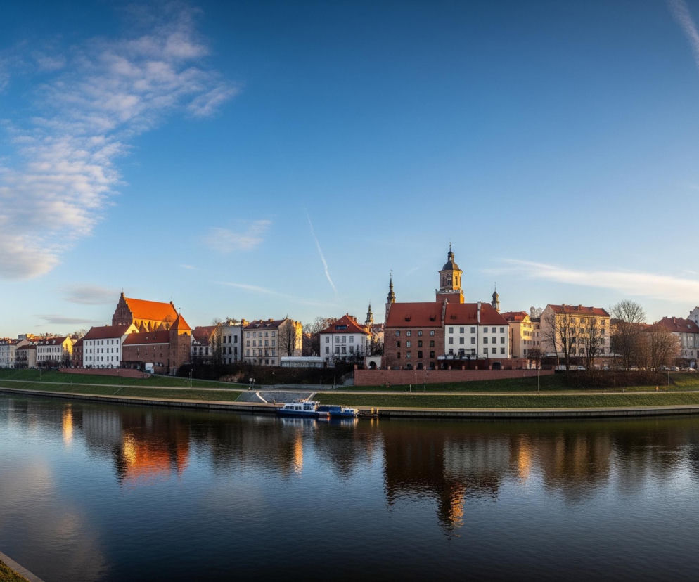 Panoramiczne ujęcie ukazuje rzekę płynącą przez miasto, oddzielając zielone, trawiaste brzegi od zabudowy miejskiej. Po prawej stronie rzeki widać szereg budynków o różnorodnej architekturze, z ceglanymi dachami w odcieniach czerwieni i pomarańczu, oraz fasadami w kolorach bieli, beżu i szarości. Wśród nich wyróżniają się budowle z wieżami, a także mniejszy statek lub barka cumujące przy prawym brzegu, którego odbicie częściowo widoczne jest na spokojnej powierzchni wody. Lewy brzeg, w dolnej części obrazu, pokryty jest zieloną trawą, a wzdłuż niego biegnie wybrukowany chodnik z czarną latarnią uliczną na pierwszym planie, podczas gdy w tle widoczny jest most o jasnej konstrukcji. Nad całym pejzażem rozciąga się bezchmurne, błękitne niebo z kilkoma delikatnymi, białymi obłokami i smugami kondensacyjnymi, oświetlone złotym światłem zachodzącego słońca, które nadaje ciepłych barw prawemu horyzontowi i niektórym budynkom.