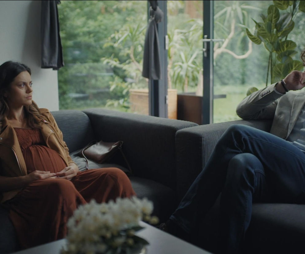 Kasia (Paulina Lasota) w zaawansowanej ciąży, siedząca zamyślona na kanapie obok Mariusza (Mateusz Mosiewicz), który wygląda na sfrustrowanego. Scena pełna napięcia w salonie, odzwierciedlająca zazdrość i narastający konflikt w serialu M jak miłość. Więcej o tym na Super Seriale.