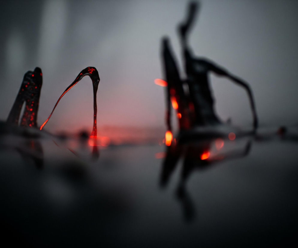 Centralnie w kadrze, z ciemnej, lśniącej powierzchni, wyłaniają się trzy abstrakcyjne, czarne formy przypominające zastygłą lawę lub smołę, z których każda zawiera jaskrawe, rozżarzone czerwone punkty i smugi. Najbardziej na pierwszym planie, po lewej stronie, widać dwie formy: jedną o nieregularnym kształcie z licznymi czerwonymi plamkami, a obok niej wygina się w łuk smukła, wydłużona struktura z intensywnie czerwoną końcówką. Na drugim planie, po prawej stronie, znajduje się większa, rozmyta forma, również z czerwonymi akcentami. Całość otacza szare tło z jaśniejszym obszarem w górnej części, natomiast dolna połowa obrazu to ciemna, refleksyjna powierzchnia.