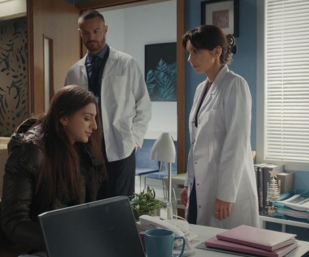 Trzy osoby w biurze lub gabinecie lekarskim: kobieta w puchowej kurtce siedzi przy laptopie, a za nią stoją kobieta i mężczyzna w białych fartuchach. Kobieta siedząca to Anka (Katarzyna Russ) w 1929 odcinku M jak miłość. Tę i inne informacje ze świata Super Seriale znajdziesz na naszym portalu.