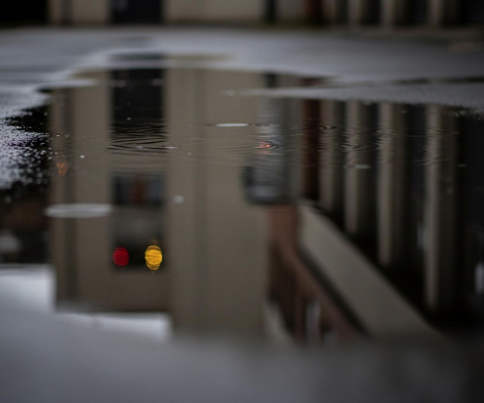 Kałuża na szarym, ziarnistym asfalcie, lekko pofałdowana od kropel deszczu, zajmuje dolną połowę kadru. Woda odbija zamazany obraz otoczenia, tworząc pionowe ciemne i jasne pasy, sprawiające wrażenie budynku z oknami. Na lewej stronie kałuży, w centralnej części, odbijają się dwa jaskrawe, okrągłe światła: jedno czerwone po lewej i jedno żółte po prawej. Górna część obrazu przedstawia rozmyte, ciemne tło, sugerujące kontynuację nawierzchni i dalsze elementy architektoniczne.