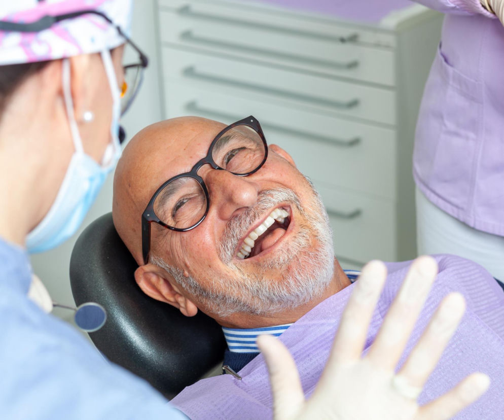 Szczęśliwy pacjent z zarostem i okularami podczas wizyty u dentystki w gabinecie stomatologicznym. Lekarka w masce medycznej i jednorazowych rękawiczkach trzyma narzędzie dentystyczne. Więcej o turystyce dentystycznej przeczytasz na Poradnik Zdrowie.