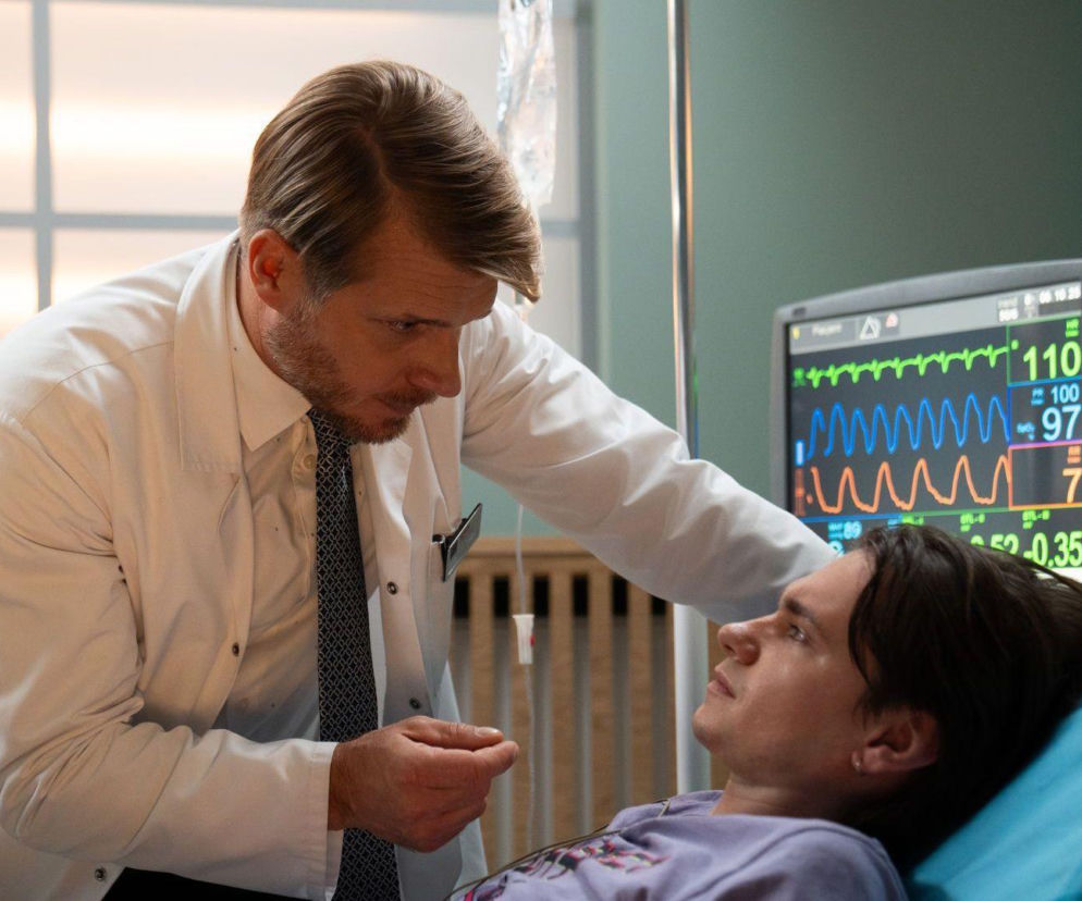 Lekarz Maks Beger (Tomasz Ciachorowski) pochyla się nad leżącym na szpitalnym łóżku Drago (Piotr Gadomski) w scenie z serialu „Na dobre i na złe”. Monitor obok pacjenta pokazuje jego parametry życiowe. Więcej informacji o serialu znajdziesz na Super Seriale.