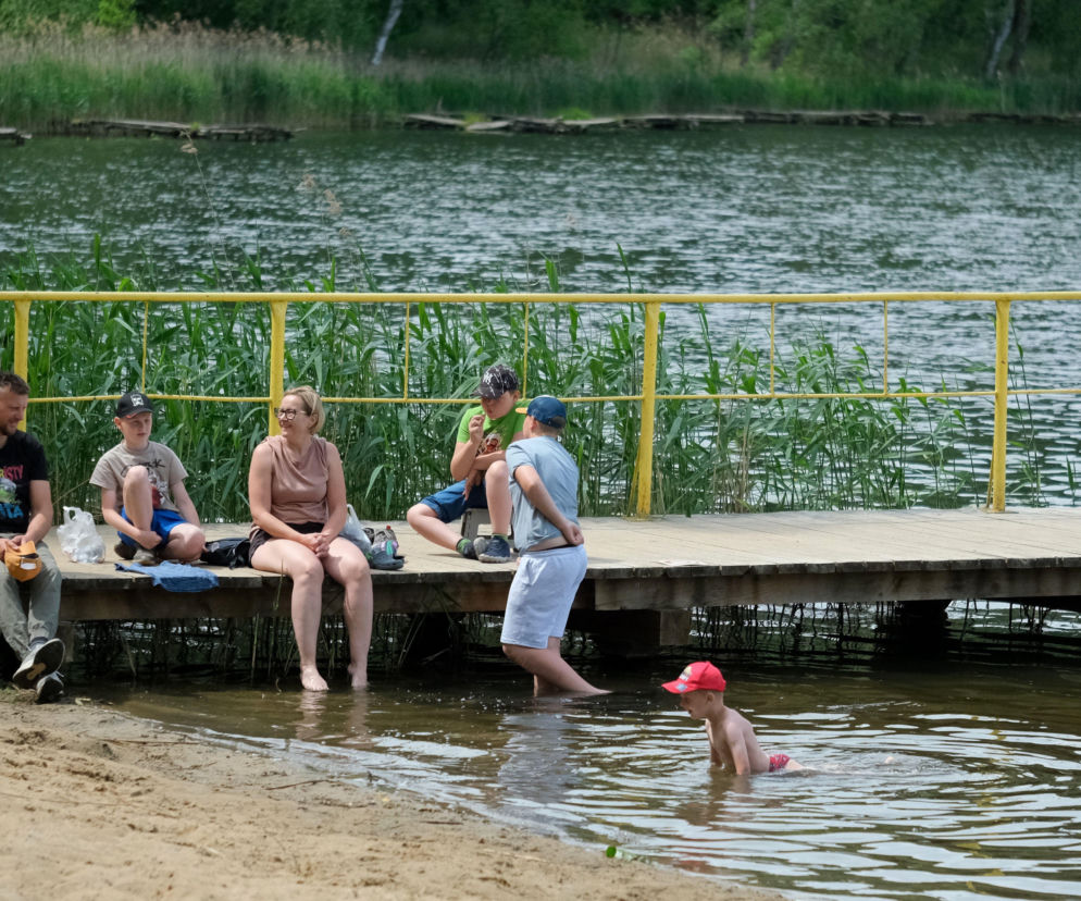 Rodzina na pomoście i w wodzie, ciesząca się weekendowym wypoczynkiem nad jeziorem w pogodny dzień. Ojciec, matka i dwójka dzieci siedzą na drewnianym pomoście, a najmłodszy chłopiec pluska się w płytkiej wodzie. Więcej o pogodzie na naszym portalu.