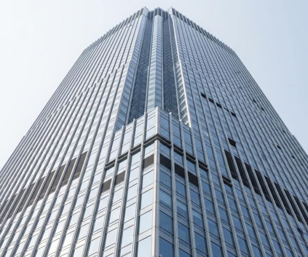 Wysoki, nowoczesny drapacz chmur o szklanej fasadzie wznosi się ukośnie ku górze, dominując centralną część kadru. Budynek charakteryzuje się regularnymi, pionowymi panelami szklanymi w odcieniach szarości i błękitu, które odbijają światło, tworząc refleksy. Dwie widoczne ściany wieżowca zbiegają się w ostrym kącie, podkreślając jego monumentalną wysokość. Tło stanowi jednolite, jasne, niemal białe niebo, które stopniowo przechodzi w delikatny błękit w prawym górnym rogu.
