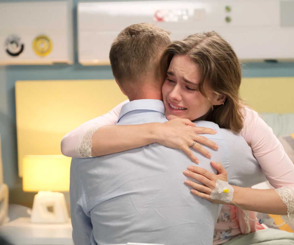 Franka (Dominika Kachlik) w różowej bluzce z koronkowymi rękawami przytula Pawła (Rafał Mroczek) w jasnoniebieskiej koszuli, płacząc. Na jej lewej ręce widoczny jest wenflon. Scena ze szpitala oddaje dramatyczne chwile przed śmiercią Franki, o której przeczytasz więcej na Super Seriale.