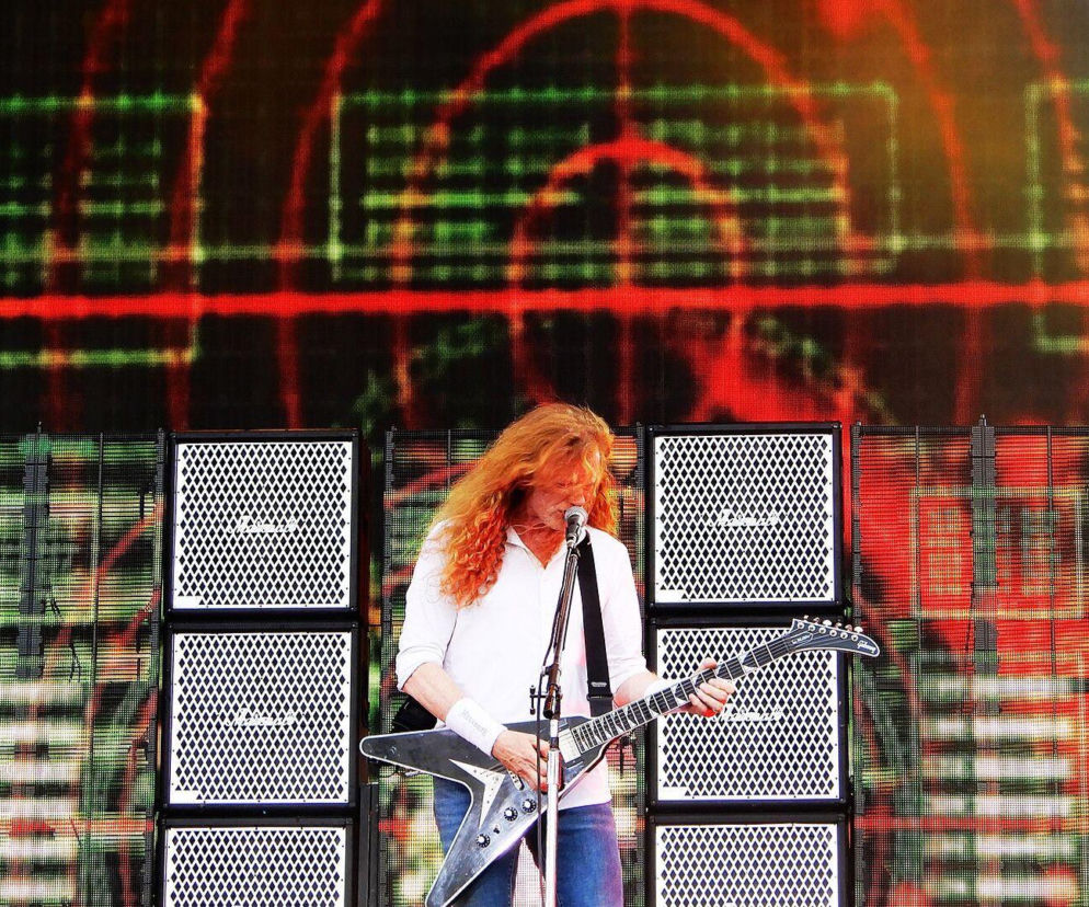 Lider Megadeth Dave Mustaine gra na gitarze na tle głośników i ekranu. O porażce albumu Risk przeczytasz na EskaRock.