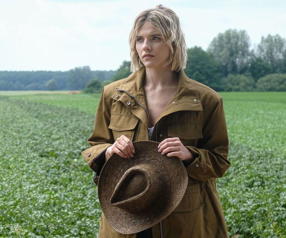 Oliwia Drożdżyk jako Janka Dąbrowska w brązowej kurtce i z kapeluszem, stoi na zielonym polu. Aktorka wcielająca się w Jankę jest jedną z bohaterek serialu Barwy szczęścia, a szczegółowe informacje na temat jej postaci znajdziesz na Super Seriale.