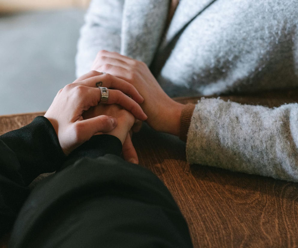 Dłonie dwóch osób, które delikatnie się dotykają, leżąc na drewnianym stole. Jedna dłoń ma na palcu pierścionek z literami, a na skórze mały tatuaż. To symbol wsparcia bliskich, niezbędnego w procesie powrotu do zdrowia po udarze mózgu i w walce z afazją, o czym przeczytasz na Poradnik Zdrowie.