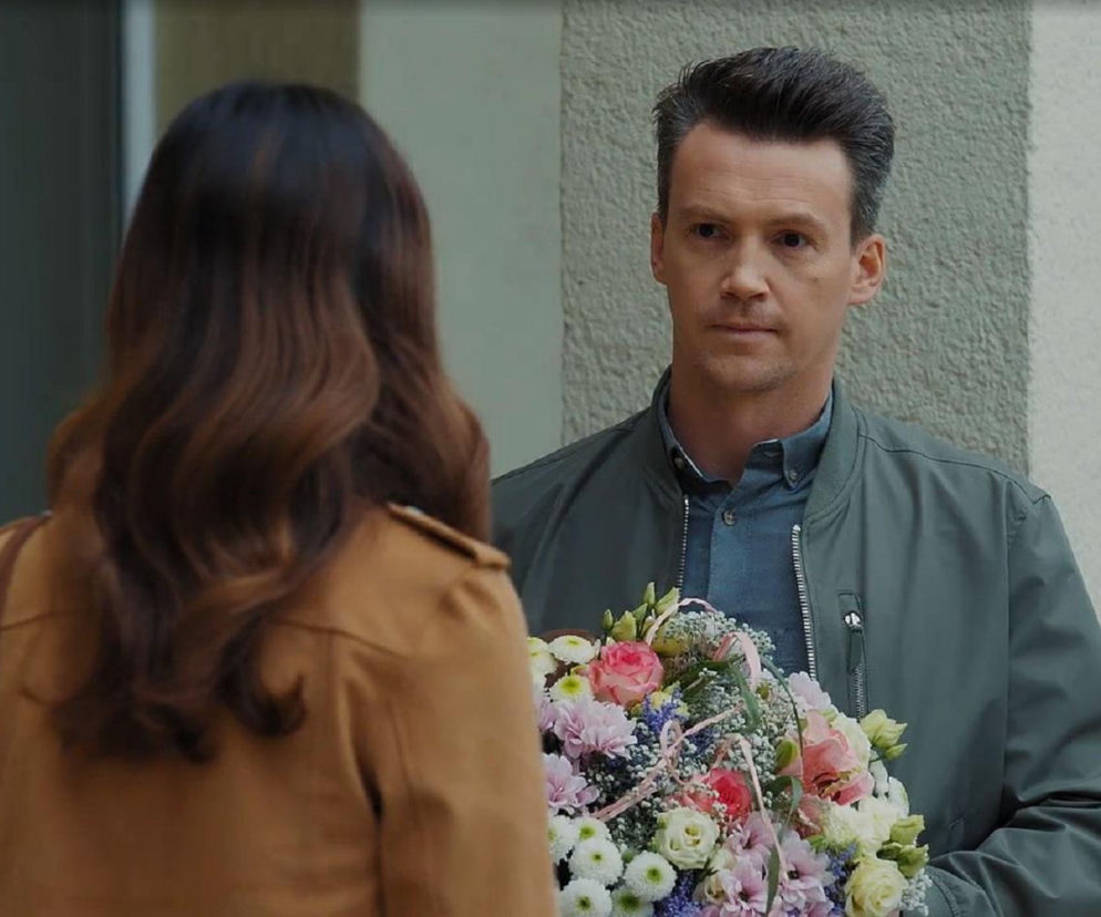 Mężczyzna trzymający bukiet kolorowych kwiatów, w tym róże i chryzantemy, rozmawia z kobietą stojącą tyłem, o długich, brązowych włosach. Zdjęcie ilustruje scenę z serialu M jak miłość, gdzie Jakub spotyka się z Kasią, o czym można przeczytać na Super Seriale.