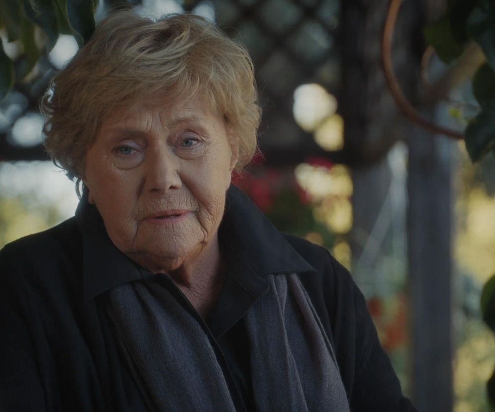 Barbara Mostowiakowa (Teresa Lipowska) w czarnej sukni i szarym szalu, wyraźnie smutna i zamyślona, co odzwierciedla jej żałobę po śmierci Franki w serialu M jak miłość. Tło stanowią niewyraźne zielone liście i konstrukcja tarasu. Więcej o losach bohaterów na Super Seriale.