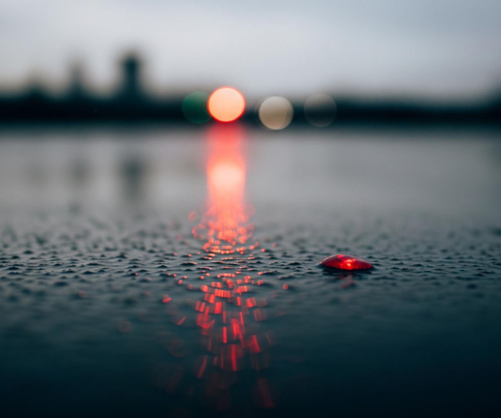 Powierzchnia pokryta kroplami wody, prawdopodobnie deszczu, jest głównym elementem obrazu, rozciągając się od samego dołu do centrum kadru. W prawym dolnym rogu znajduje się pojedyncza, intensywnie czerwona kropla wody, która jest wyraźnie widoczna i odbija światło. Od niej, poprzez liczne mniejsze krople, rozciąga się w głąb obrazu intensywna, czerwona smuga światła, będąca odbiciem większego, rozmytego źródła światła, prawdopodobnie latarni lub sygnalizacji, znajdującego się w oddali. Tło jest rozmyte, z widocznymi, niewyraźnymi światłami bokeh w kolorach czerwonym, zielonym i białym, a także z ciemnymi, zamazanymi sylwetkami budynków na horyzoncie.