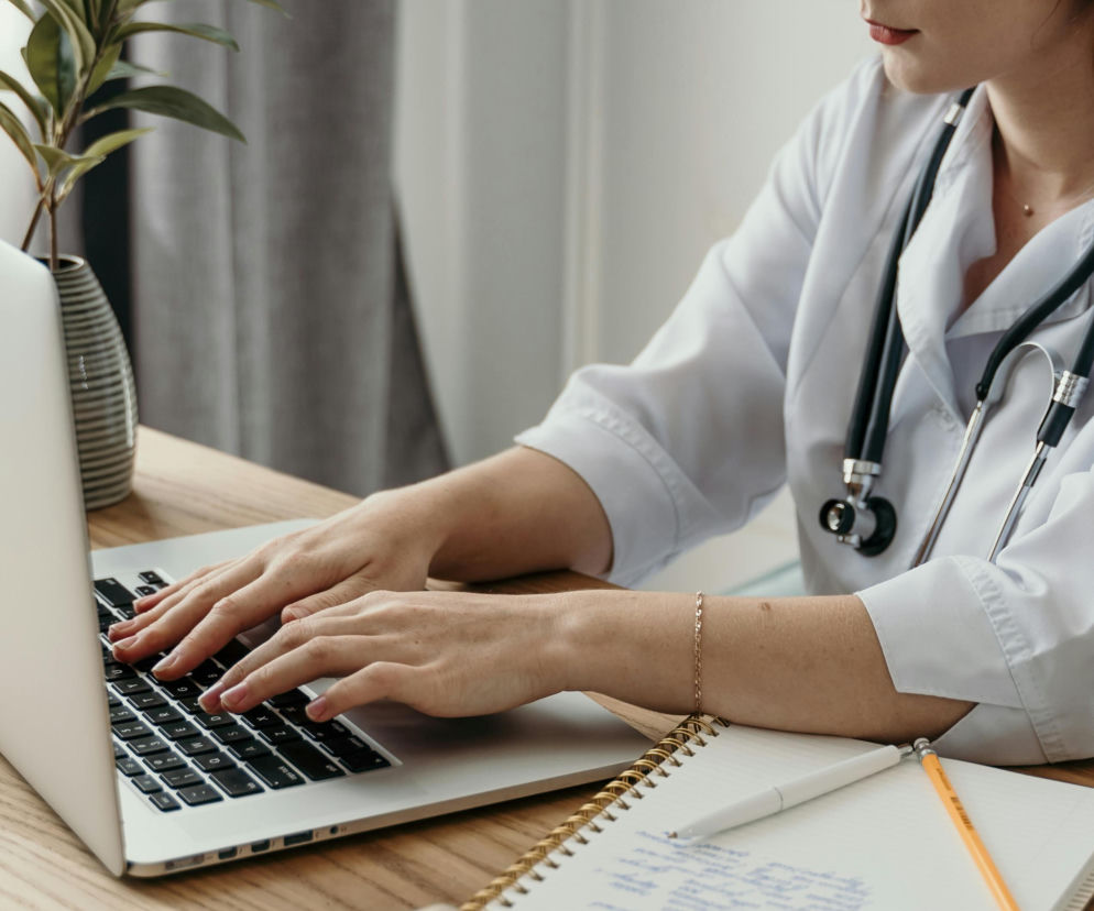 Lekarka w białym kitlu ze stetoskopem na szyi, pisząca na laptopie, obok notes i książki. Zdjęcie symbolizuje rozwój e-zdrowia i sztucznej inteligencji w medycynie, o czym można przeczytać na Super Biznes.