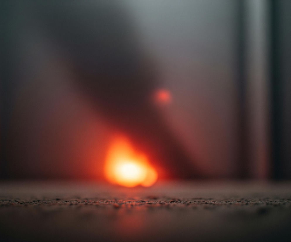 Pośrodku obrazu dominuje rozmyta, jasnopomarańczowa poświata, przypominająca płomień lub intensywne źródło światła. Otacza ją ciemne tło w odcieniach szarości i brązu, które staje się jaśniejsze i cieplejsze w pobliżu źródła światła, a ciemniejsze i chłodniejsze w górnych rogach oraz po bokach. Na pierwszym planie widoczna jest szczegółowa, ziarnista powierzchnia, która jest ostra i odzwierciedla subtelnie pomarańczowe światło, tworząc poziome odbicia.