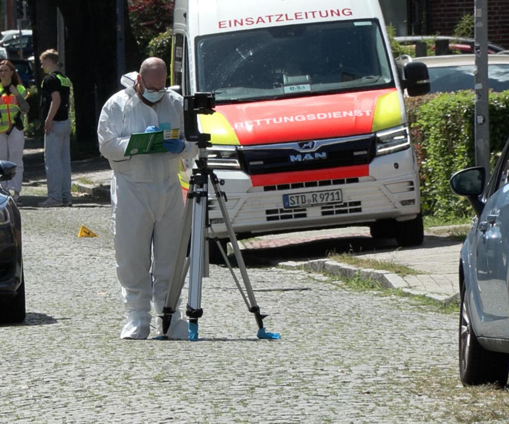 Funkcjonariusz w białym kombinezonie ochronnym i masce bada miejsce strzelaniny w Stade, gdzie zginęło pięć osób. Obok niego widoczny statyw fotograficzny i radiowóz, a w tle inne służby. Więcej o tragedii w Hamburgu przeczytasz na naszym portalu.