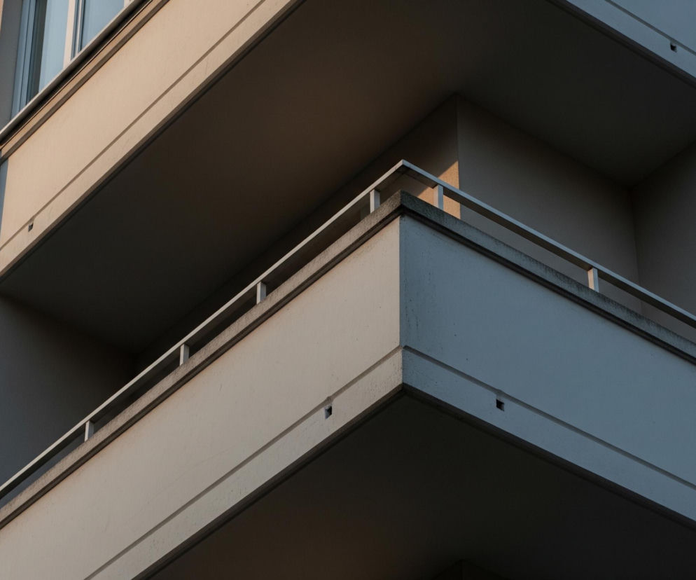 Fragment nowoczesnego budynku mieszkalnego, przedstawiający kilka balkonów i część elewacji. Dominują jasne, neutralne odcienie beżu i szarości, z kontrastującymi cieniami rzucanymi przez wystające elementy architektoniczne. Na ścianach widać tynk o drobnej, ziarnistej fakturze oraz prostokątne okna z białymi ramami, których szyby odbijają światło. Balustrady balkonów są wykonane z poziomych, metalowych prętów w jasnym kolorze.