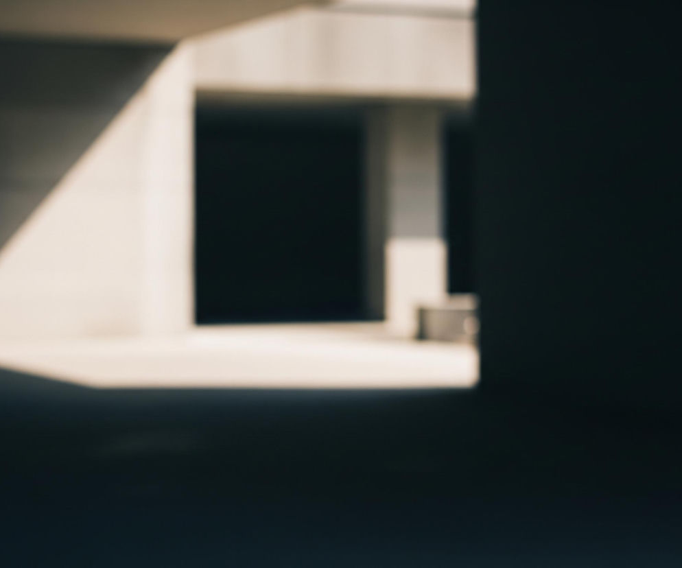 Rozmyty obraz przedstawia wnętrze nowoczesnego budynku, z dominującymi ciemnymi i jasnymi odcieniami. Po prawej stronie widać ciemną, pionową powierzchnię, która zasłania część widoku. W centrum i po lewej stronie obrazu dominuje jasna, beżowa przestrzeń, oświetlona skośnym światłem tworzącym geometryczne kształty. Architektura charakteryzuje się prostymi liniami i kątami, z widocznymi filarami i prostopadłościennymi otworami, prowadzącymi w głąb ciemniejszych partii konstrukcji.