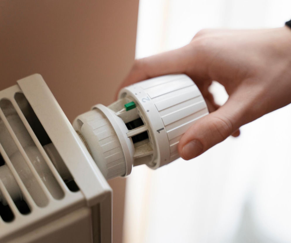 Dłoń osoby przekręcającej białą głowicę termostatyczną przy grzejniku. O przeglądzie przed sezonem przeczytasz w Muratordom.