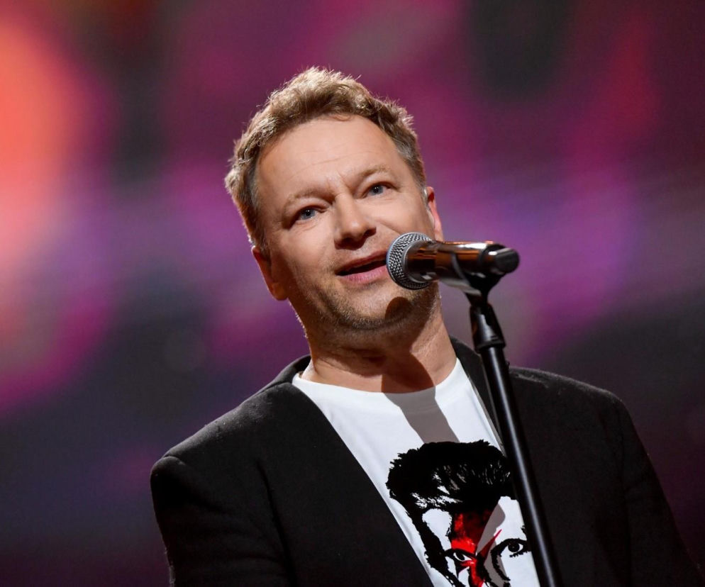 Maciej Stuhr w czarnej marynarce i białej koszulce z wizerunkiem Davida Bowiego, przemawiający do mikrofonu na scenie z rozmazanym, kolorowym tłem. Aktor o krótkich włosach i lekkim zaroście patrzy w górę. Więcej o metamorfozie Macieja Stuhra przeczytasz na naszym portalu.